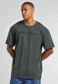 Dark green cotton t-shirt with a relaxed fit, featuring a black script logo across the chest and short sleeves. Simple neckline design.