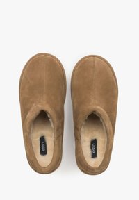 Een paar bruine suède slippers met eenplush crèmekleurige binnenvoering en zwarte labels aan de binnenkant, van bovenaf weergegeven op een witte achtergrond.