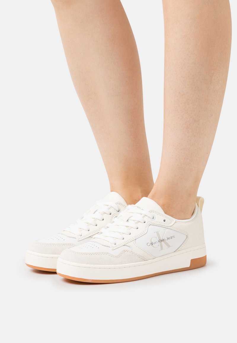 Paire de baskets blanches Calvin Klein Jeans avec des accents en daim beige et des semelles en gomme, portées pieds nus sur fond blanc.