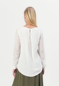 Weiße Bluse mit strukturierten, gemusterten langen Ärmeln und einem Rückenausschnitt im Schlüsselaugen-Stil. Weicher Stoff mit abgerundetem Saum und leicht lockerer Passform.