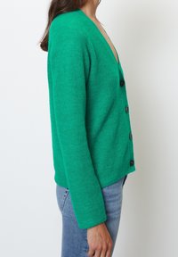 Cárdigan de punto verde con escote en V profundo, mangas largas y cinco botones negros, con una textura de tela y un ajuste ligeramente holgado.