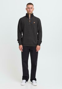 Mørkegrå genser med kvart glidelås, ribbede mansjetter og kant. Har et lite lærlogo-merke. Kombinert med svarte bukser og hvite sneakers.