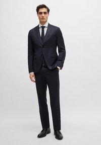 Marineblauer Anzug mit einreihiger Jacke, Reverskragen und zwei Fronttaschen. Maßgeschneiderte Hose mit glatter Oberfläche. Schwarze Schuhe inklusive.