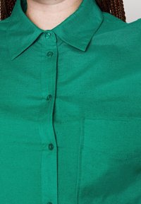 Grön skjorta med knappar i slätt tyg, med krage, sex knappar fram och en liten bröstficka.