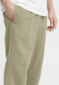 Helle olivgrüne Hose aus strukturiertem Stoff, mit Seitentaschen und einer lockeren Passform. Sichtbare Nähte betonen das Design.