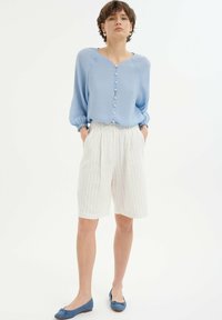 Hellbluse mit langen Ärmeln, Knopfleiste und gerafften Manschetten, kombiniert mit weißen Shorts mit feinen Streifen. Blaue flache Schuhe runden das Outfit ab.