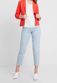 Czerwony sweter w formie marynarki na białym polo, w połączeniu z jasnoniebieskimi, skróconymi dżinsami oraz białymi sandałami na obcasie. Gładkie faktury i prosty design.