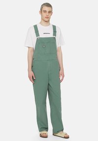 Green cotton overalls with adjustable straps, front pockets, and side buttons, paired with a white T-shirt featuring black text.
