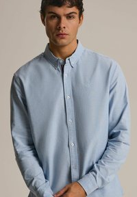 Jasnoniebieska koszula z guzikami, z kołnierzykiem, długimi rękawami i subtelną teksturą. Posiada małe logo na piersi i białe guziki.