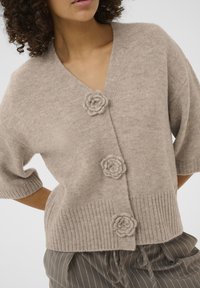 Beige strikket cardigan med V-hals, korte ærmer og tre dekorative knapper formet som blomster. Ribstrikket kant giver tekstur.