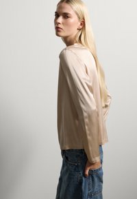 Top lungo a maniche di satin beige chiaro con una vestibilità morbida, scollo rotondo e texture liscia, abbinato a jeans in denim blu a vita alta.