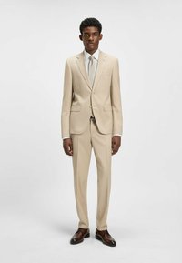 Costume beige avec une veste à boutons simples, revers crantés et un pantalon assorti. Accompagné d'une cravate de couleur claire et de chaussures brunes.