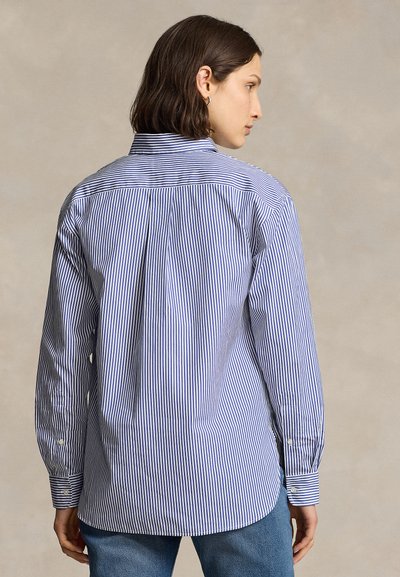 Persona joven con cabello corto y oscuro, vestida con una camisa de rayas verticales azules y blancas con botones y vaqueros azules mirando hacia atrás.