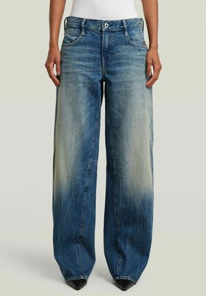 Person trägt locker sitzende, blau ausgewaschene Jeans mit weitem Bein und schwarze spitze Schuhe und steht vor einem schlichten Hintergrund.