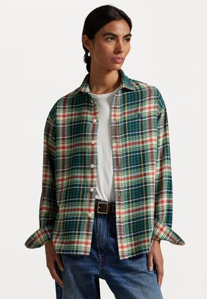 Donna che indossa una camicia a quadri verde, blu navy, rossa e bianca, sopra una maglietta bianca, abbinata a jeans blu e una cintura nera con fibbia dorata.