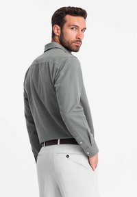 Chemise grise à manches longues avec un col boutonné, texture lisse et design ajusté, associée à un pantalon gris clair et une ceinture noire.