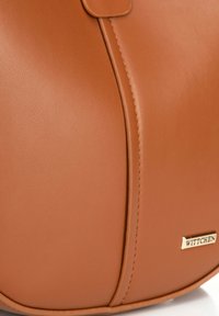Borsa a mano in pelle marrone con texture liscia e cuciture a vista, dotata di un'etichetta del marchio in tonalità oro. Forma arrotondata con un design elegante.