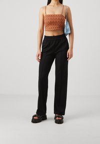 Avklippt orange topp med vågmönster, svarta byxor med elastisk midja och kraftiga svarta sandaler med remmar och dekorativa detaljer.