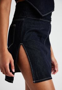 Sötétkék farmer szoknya oldalsó sliccel, fehér varrással és testre szabott derékrésszel. Texturált anyag sima felülettel.