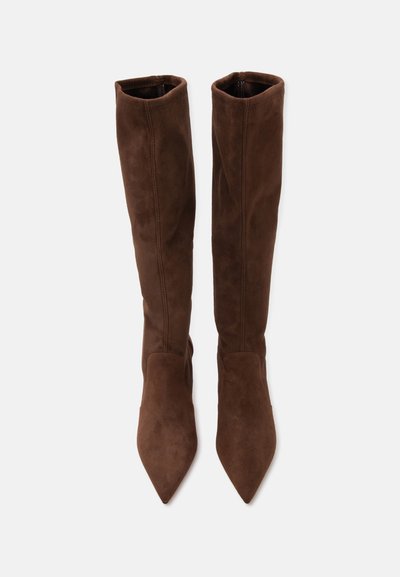 Paire de bottes hautes en daim marron avec bouts pointus, debout sur un fond clair uni.