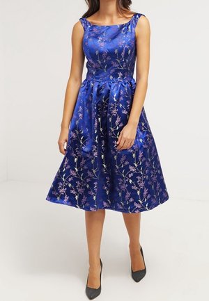 Mujer con un vestido de satén azul real, sin mangas, hasta la rodilla, con estampados florales rosas y tacones negros de punta.