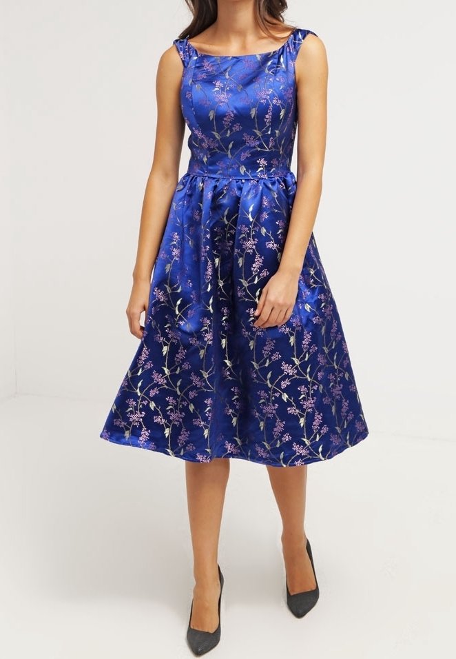 Femme portant une robe en satin bleu roi sans manches, longueur genou, avec des motifs floraux roses et des talons noirs à bout pointu.
