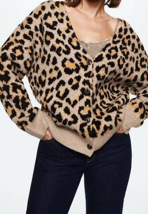 Vrouw met een beige leopardprint vest met knopen over een taupekleurig hemdje met kant en donkerblauwe spijkerbroek, met haar handen in de zakken.