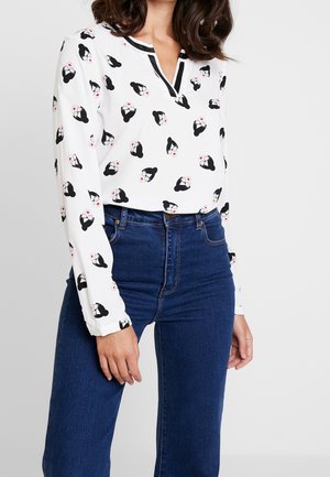 Femme portant un chemisier blanc à manches longues avec un motif stylisé de visages féminins noir et rouge, assorti à un jean taille haute bleu foncé.
