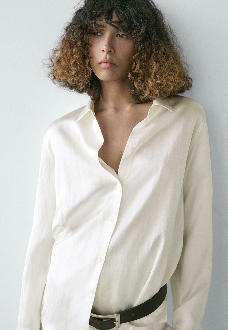 Jonge persoon met krullend ombre haar, gekleed in een oversized wit overhemd met knopen en een zwarte riem, staand tegen een effen lichte achtergrond.