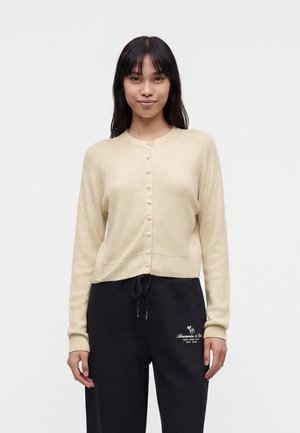 Junge Frau mit glatten dunklen Haaren, die eine beige, geknöpfte Strickjacke und schwarze Hose mit weißem Logo am linken Oberschenkel trägt.