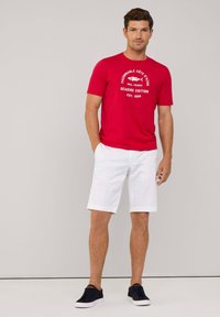 Camiseta de algodón roja con estampado gráfico en blanco, combinada con pantalones cortos de algodón blancos. Zapatillas negras completan el atuendo. Diseño simple y casual.
