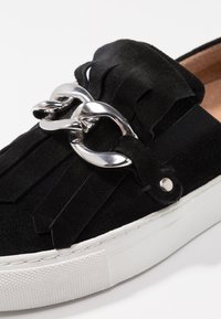 Chaussure à enfiler en daim noir avec des détails à franges, un accent en chaîne argentée et une semelle en caoutchouc blanche avec une adhérence texturée.