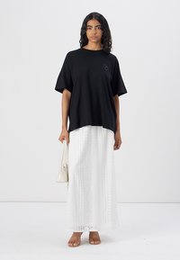 Sort oversized t-shirt med korte ærmer, parret med en hvid maxiskirt med blonder. Modellen holder en lille hvid handbag.