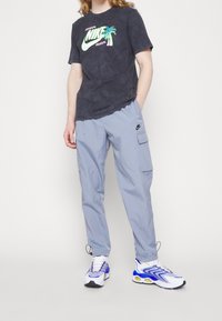 Černé grafické tričko s palmovým vzorem, šedé cargo kalhoty s kapsami a bílé tenisky s modrými akcenty a texturovanou síťovinou.