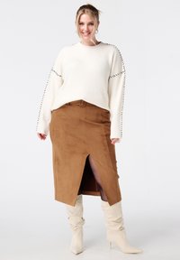 Glunderende vrouw met blond haar in een crèmekleurige trui, een bruine rok met riem en een split aan de voorkant, zwarte panty en crèmekleurige hoge hakken op een witte achtergrond.