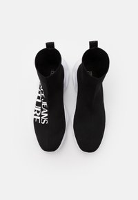 Zapatos de punto negros hasta el tobillo con suelas blancas que presentan texto impreso en el lado exterior. Textura suave y diseño elástico similar a un calcetín.