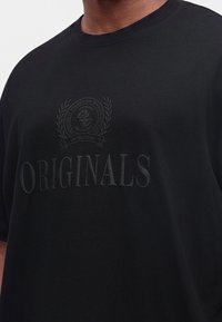 Μαύρο μπλουζάκι με ανάγλυφο κείμενο "ORIGINALS" και κυκλικό έμβλημα από πάνω με φύλλα δάφνης και μικρές λεπτομέρειες κειμένου.