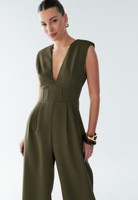 Olivově zelený overal s hlubokým výstřihem do V, krátkými rukávy a řaseným pasem. Hladká látka a široké nohavice zvýrazňují siluetu. Zlaté doplňky.