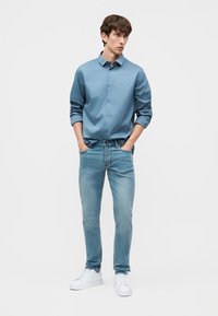 Νέος άνδρας στέκεται με τα χέρια στις τσέπες, φορώντας μπλε πουκάμισο με κουμπιά, ανοιχτόχρωμα τζιν και λευκά αθλητικά παπούτσια, πάνω σε λευκό φόντο.