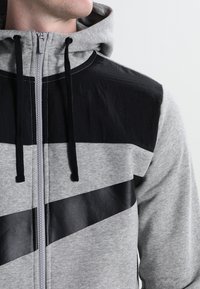 Muž oblečený v šedé mikině na zip s černými diagonálními detaily a černými šňůrkami, viditelný od krku po střed trupu na jednotném pozadí.