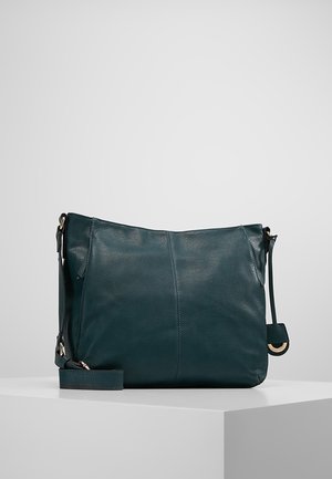 Sac à bandoulière en cuir vert foncé avec sangle réglable exposé sur un piédestal blanc sur fond gris.