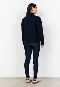 Navy fleece-pullari korkealla kauluksella, kohokuvioitu pinta, pitkät hihat, yhdistettynä tummiin farkkuihin ja vaaleanpunaiseen kenkäpariin.