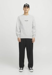 Sweatshirt gris à col ras du cou avec logo en texte noir, associé à un pantalon noir ample et des baskets noires, le tout sur un fond blanc uni.