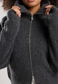 Sweater de color gris carbón con cremallera, textura acanalada, alto cuello, cremallera prominente y mangas anchas. El material parece cálido y ligero.