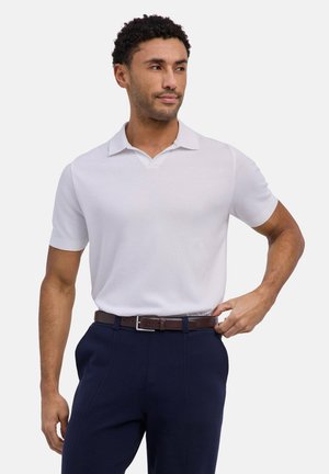 Uomo che sistema una cintura marrone indossando una polo bianca a maniche corte e pantaloni blu navy su sfondo bianco uniforme.