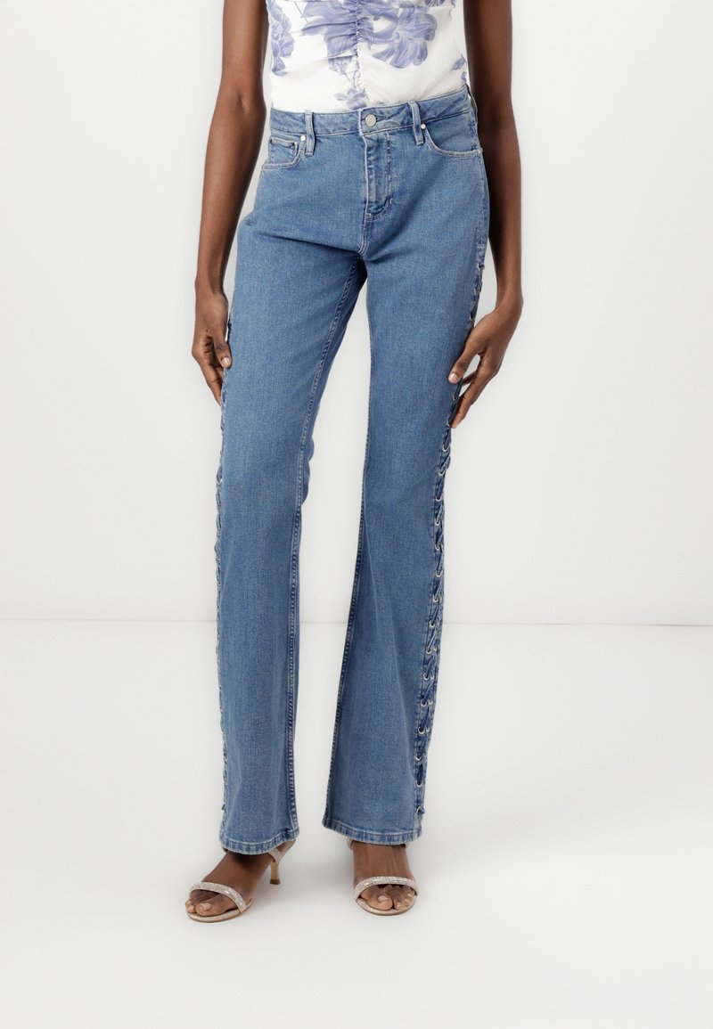 Femme portant un jean en denim bleu clair avec des détails à lacets le long des coutures extérieures, associé à des sandales à talons à brides et un haut à imprimé floral.