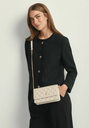 Kobieta ubrana w czarną rozpinaną kurtkę i czarne spodnie, niosąca beżową pikowaną torebkę na ramię z złotym łańcuszkiem.