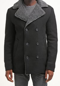 Svart dubbelknäppt rock med en grå shearling-krage, utrustad med sidofickor och knäppningar, tillverkad av ett texturerat tyg.