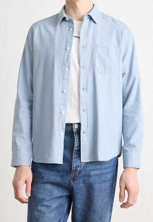 Man wearing light blue button-up shirt over white graphic t-shirt and blue jeans, standing against plain background.