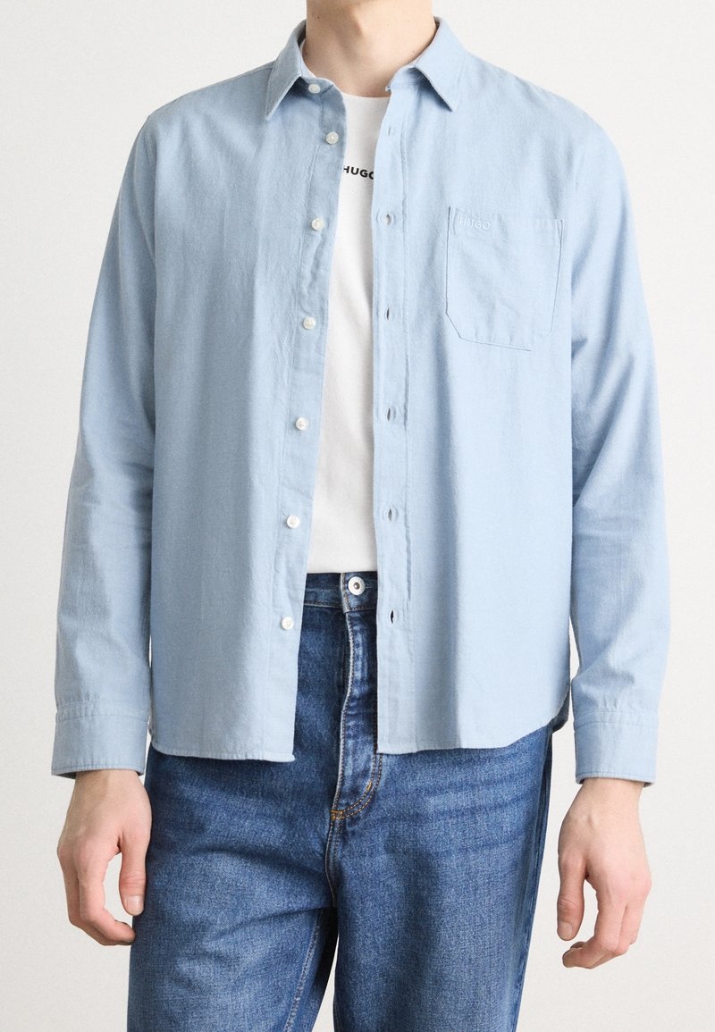 Homme portant une chemise bleu clair boutonnée par-dessus un t-shirt graphique blanc et un jean bleu, debout devant un fond uni.